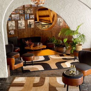 Sfeervolle zithoek met het Elitis vloerkleed Penny Lane, omringd door planten, foto's aan de muur en retro meubels.