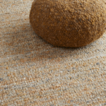 Beige Perletta vloerkleed Gravel met daarop een bruin, geweven poef.
