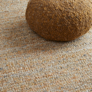 Beige Perletta vloerkleed Gravel met daarop een bruin, geweven poef.