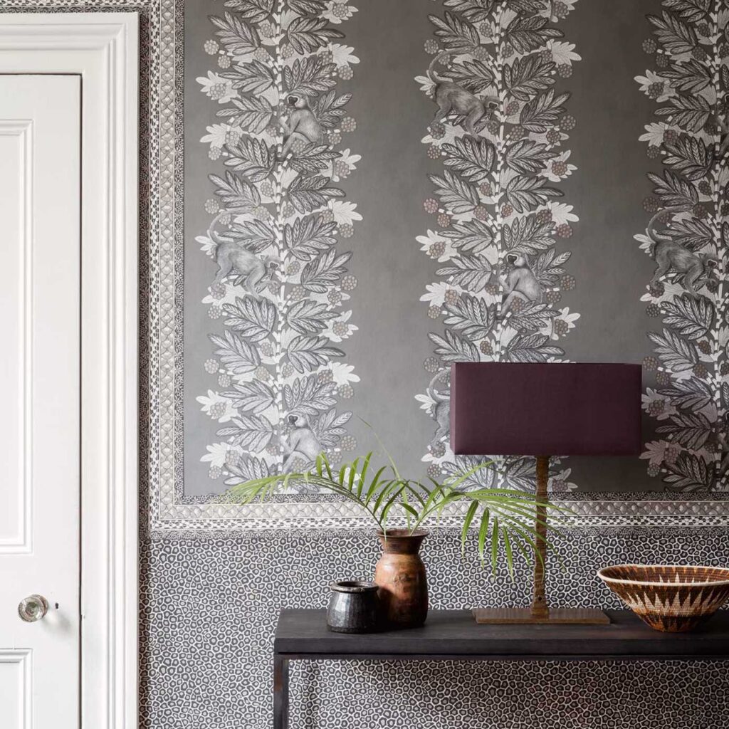 Grijze Ardmore Acacia-behang van Cole and Son met illustraties van apen, bladeren en bessen, gecombineerd met een donkere console en decoratieve objecten.