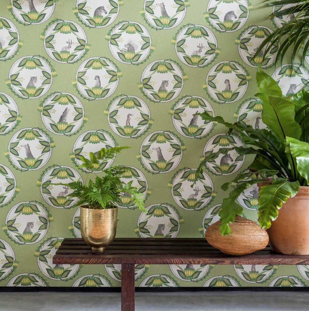Groen Ardmore Cameos-behang van Cole & Son met Afrikaanse dierportretten en grote tropische kamerplanten op een houten bank.