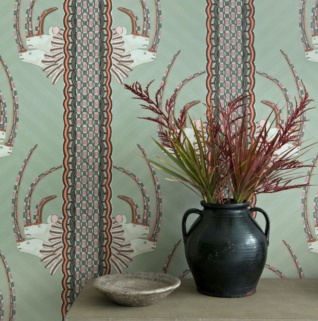 Behang met het Jabu-patroon van Cole and Son, featuring gestileerde antilopekoppen en kleurrijke geometrische details, met een zwarte vaas met siergrassen ervoor.