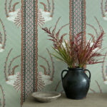 Behang met het Jabu-patroon van Cole and Son, featuring gestileerde antilopekoppen en kleurrijke geometrische details, met een zwarte vaas met siergrassen ervoor.
