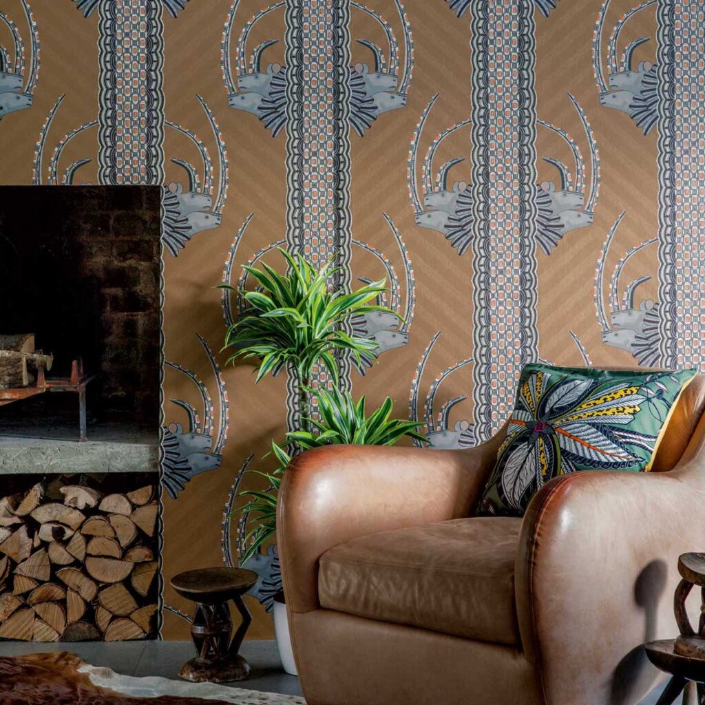 Leren fauteuil met kleurrijk kussen voor Ardmore Jabu-behang van Cole and Son met grafisch patroon van antilopekoppen en kralen.