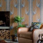 Leren fauteuil met kleurrijk kussen voor Ardmore Jabu-behang van Cole and Son met grafisch patroon van antilopekoppen en kralen.