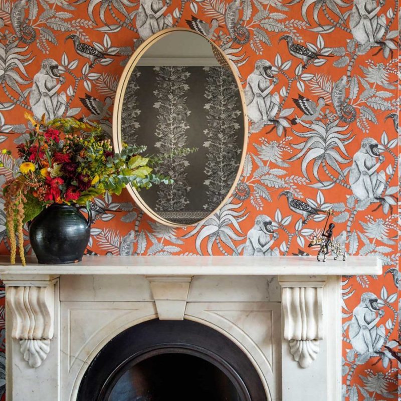 Marmeren schouw met een kleurrijk bloemstuk en een ovaal spiegel tegen een oranje Ardmore Savuti-behang met exotische dieren en planten.