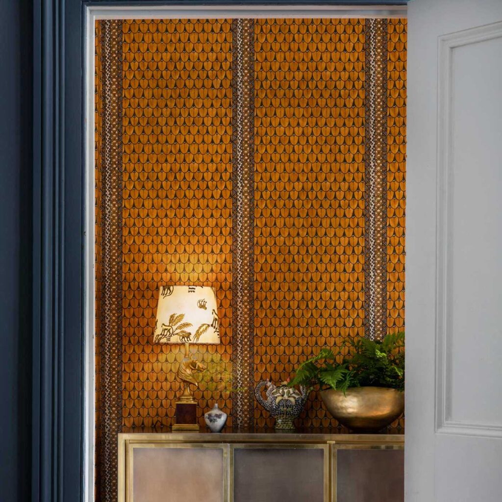 Oranje muur met het Cole and Son Ardmore Border 109/5026 behang, versierd met een gouden kast, lamp en planten.