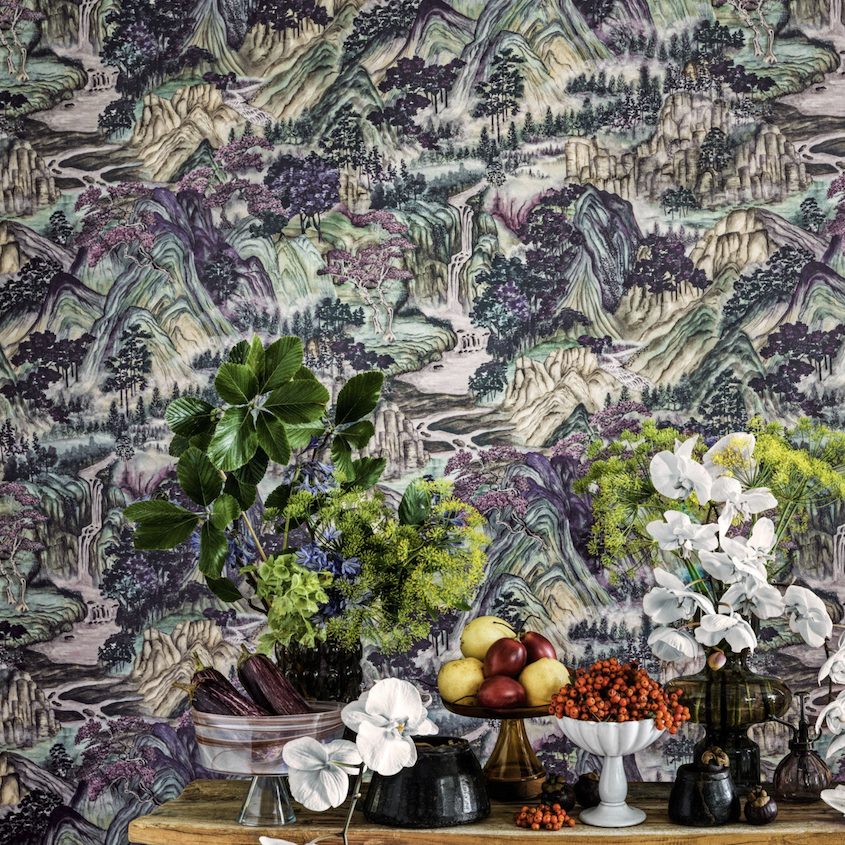 Stilleven met bloemen en fruit op een houten tafel tegen Cole and Son Mountainscape-behang met een schilderachtig berglandschap.
