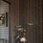 Donkere behangwand met het Cole and Son Woods & Pears-patroon van bomen en peren, naast een geometrische muur en een vaas met bloemen.