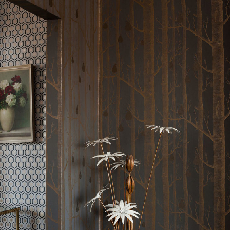 Donkere behangwand met het Cole and Son Woods & Pears-patroon van bomen en peren, naast een geometrische muur en een vaas met bloemen.