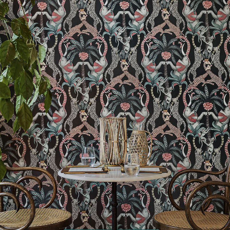 Sfeervolle interieurhoek met een ronde tafel en rotan stoelen tegen het kleurrijke Cole and Son Ardmore Jabula Safari Totem behang.