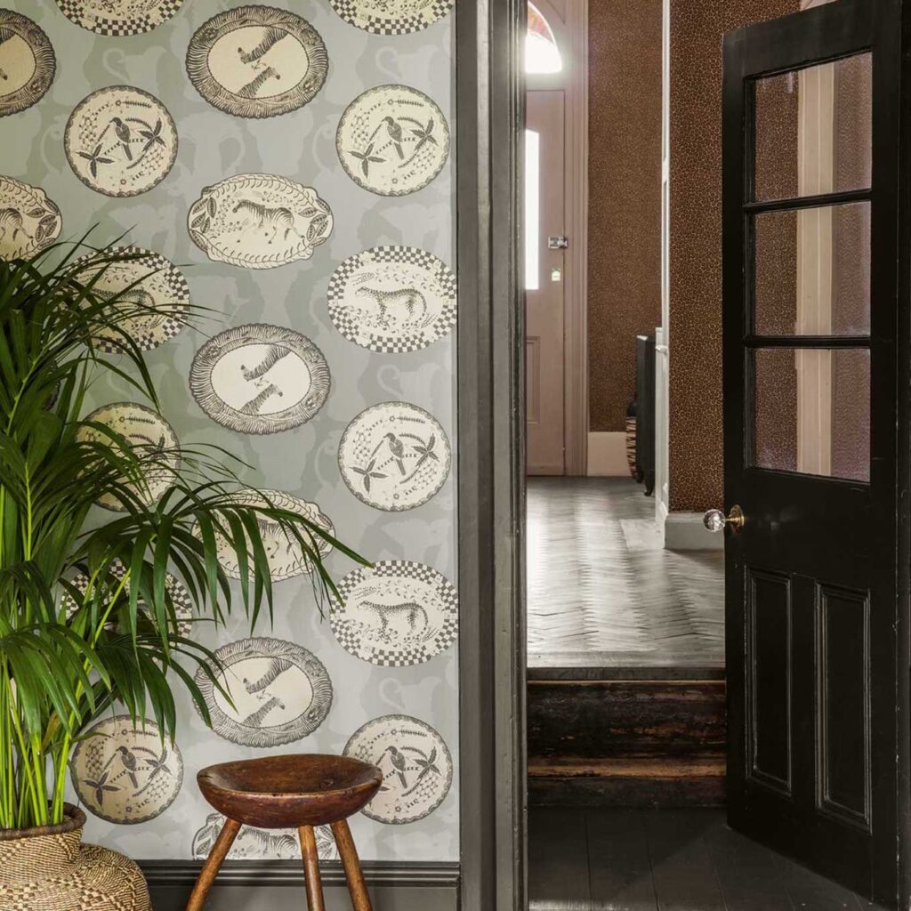 Wand met Cole and Son Ardmore Matrinah behang met decoratieve dierenprints in een sfeervol interieur met houten kruk en groene plant.