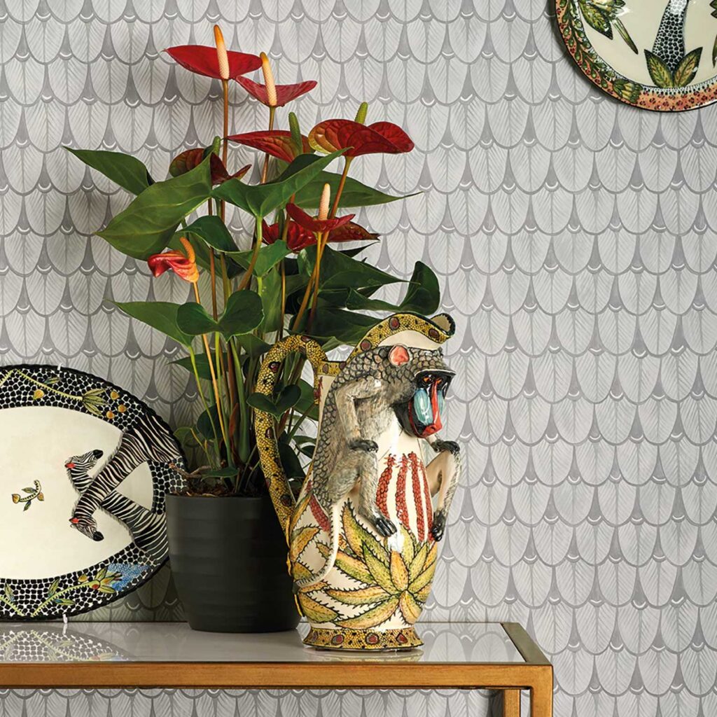 Decoratieve opstelling met Ardmore Narina behang van Cole and Son, een kleurrijke aap-vormige kan, een zwarte bloempot met rode anthuriums en keramisch servies.