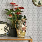 Decoratieve opstelling met Ardmore Narina behang van Cole and Son, een kleurrijke aap-vormige kan, een zwarte bloempot met rode anthuriums en keramisch servies.