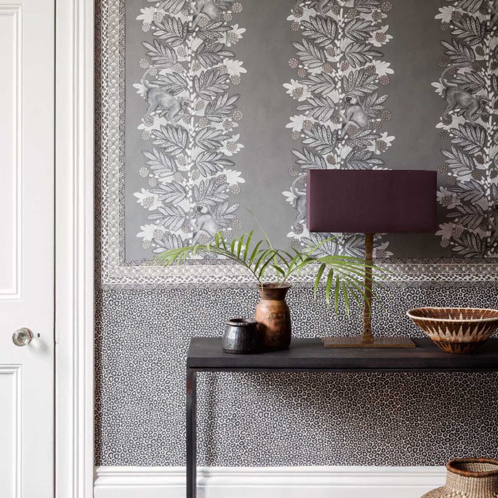 Grijze wand met Cole and Son Ardmore Senzo Spot behang, gedecoreerd met grafische bladeren, apen en een zwarte tafel met aardewerk en een lamp.