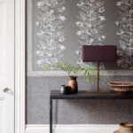 Grijze wand met Cole and Son Ardmore Senzo Spot behang, gedecoreerd met grafische bladeren, apen en een zwarte tafel met aardewerk en een lamp.