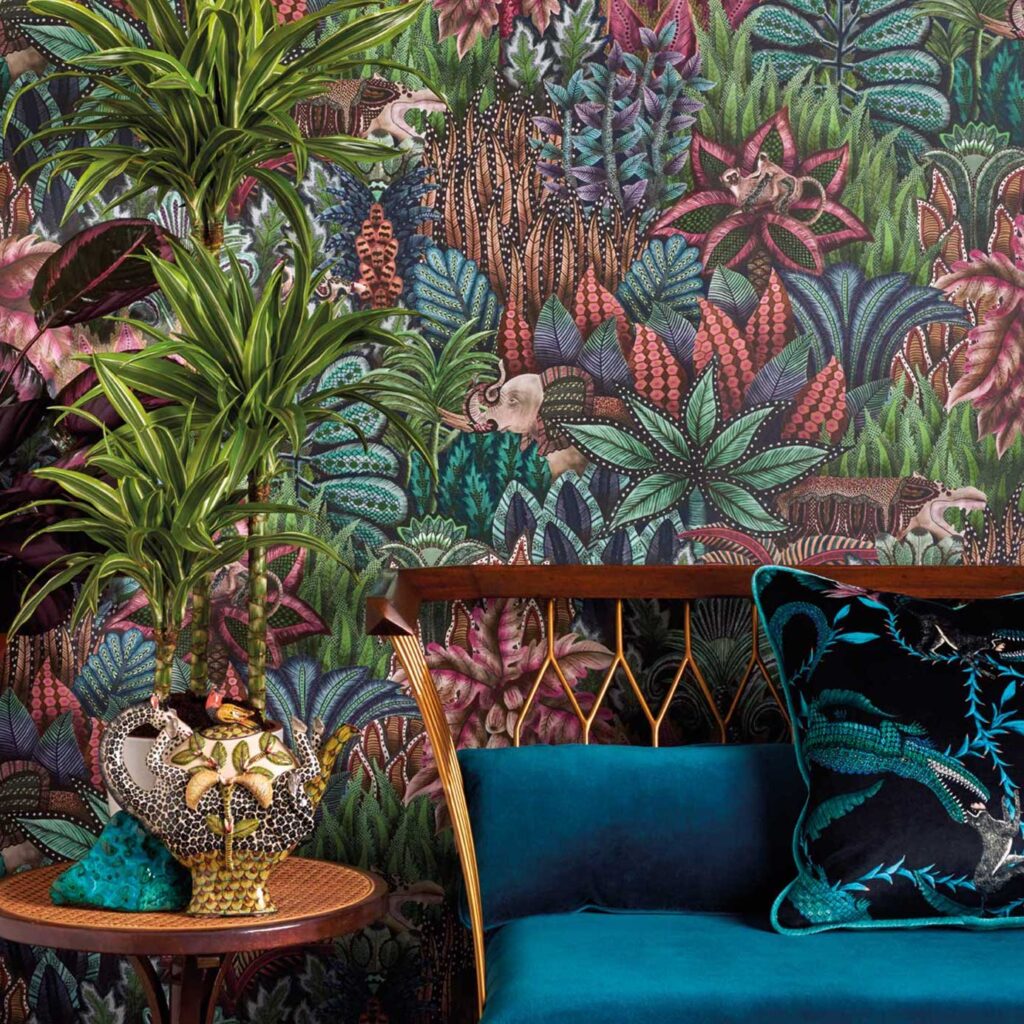 Behang Ardmore Singita van Cole and Son met kleurrijk jungle-patroon, omringd door planten en een fluwelen bank met sierkussen.