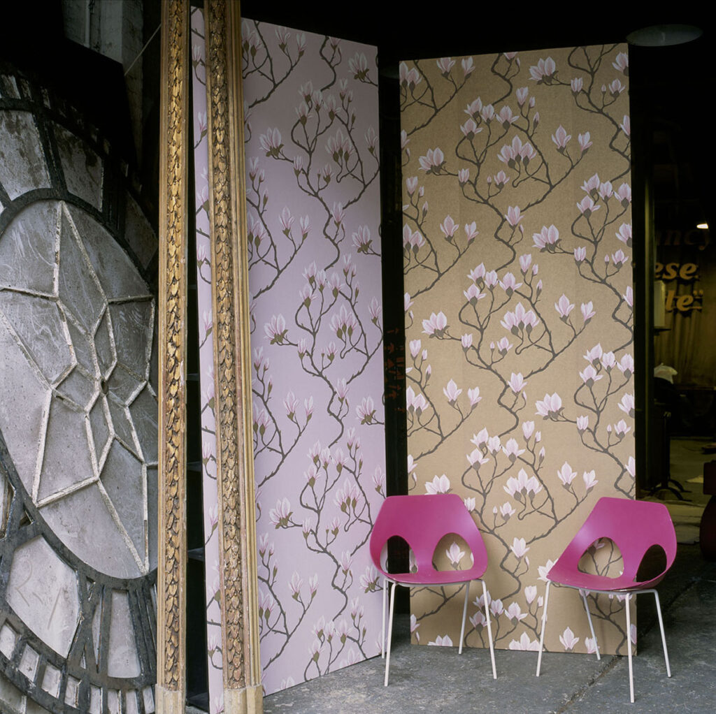 Twee panelen met Cole and Son Magnolia-behang in roze en beige tonen een bloemmotief, met daarvoor twee roze designstoelen.
