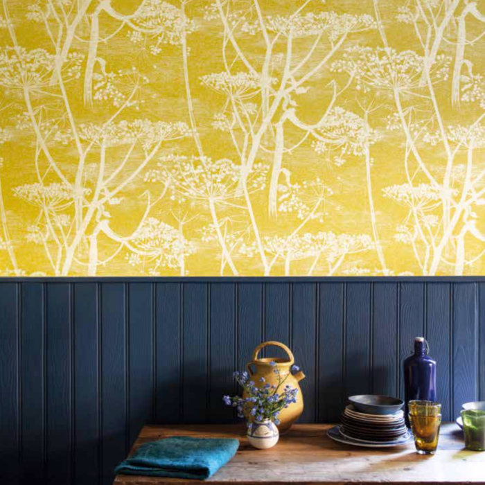 Geel behang met wit Cow Parsley patroon van Cole and Son uit de Contemporary Collection, boven een donkerblauw houten lambrisering en een rustiek houten tafelblad.