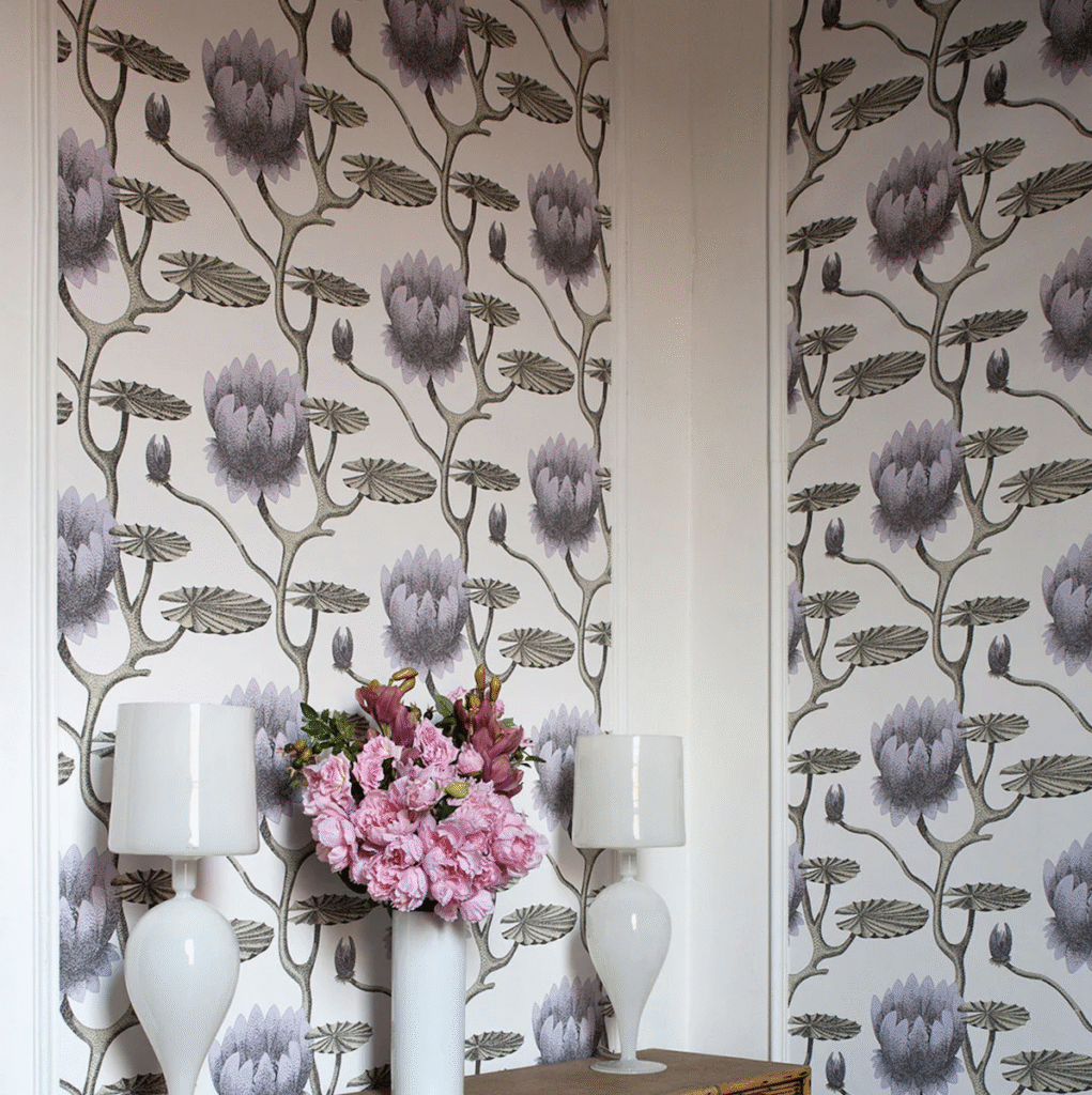Behang met het Summer Lily-patroon van Cole and Son in een sfeervolle interieursetting met een houten kastje, witte lampen en een vaas met roze bloemen.