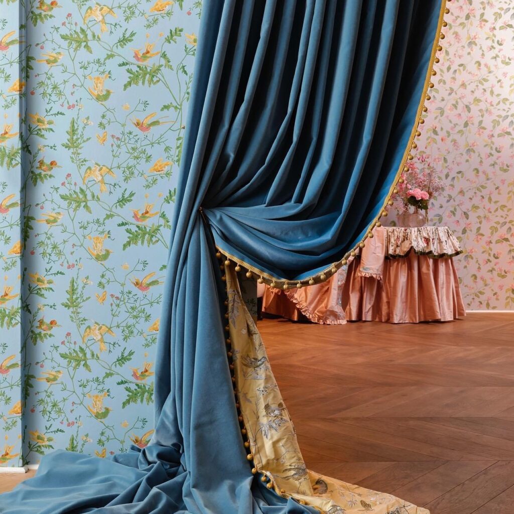 Blauw Cole & Son Hummingbirds-behang met kleurrijke vogels en bloemen, deels bedekt door een elegante blauwe fluwelen gordijn.