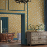 Sfeervolle kamer met Cole & Son Hummingbirds-behang in mosterdgeel, een koperen badkuip en vintage houten ladekast met bloemen.