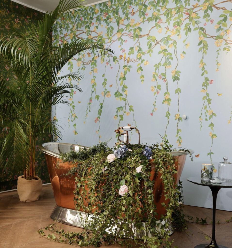 Koperen badkuip met bloemen en klimop in een botanisch interieur met Cole & Son Hummingbirds Flora-behang.