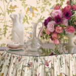 Sfeervolle setting met een ronde tafel bekleed met Cole & Son Hummingbirds-stof, omringd door bloemen, keramiek en een glazen karaf.