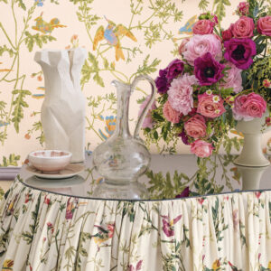 Sfeervolle setting met een ronde tafel bekleed met Cole & Son Hummingbirds-stof, omringd door bloemen, keramiek en een glazen karaf.
