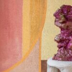 Golvende, abstracte sculptuur in roze en paarstinten op sokkel, voor kleurrijk geometrisch schilderij uit de serie "Éclats Cosmiques" van Véronique de Soultrait.