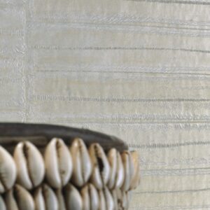 Detail van Elitis-Anguille-8 behang met een glanzende textuur in lichte tinten en een decoratieve schaal met kaurischelpen op de voorgrond.