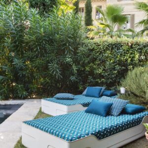 Twee moderne ligbedden met Elitis Aquarama outdoor stof en blauwe kussens staan in een weelderige tuin.