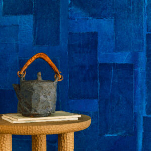 Ronde houten bijzettafel met decoratieve kan en boeken tegen een blauw Elitis Amami RM 1035 05 behang.