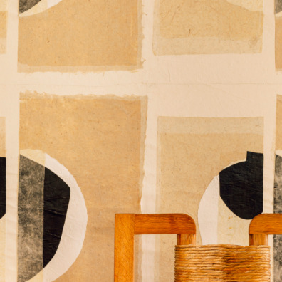 Detail van het Elitis Izu RM 1030 02 behang met abstracte geometrische patronen in aardetinten, met op de voorgrond een houten stoel met gevlochten zitting.