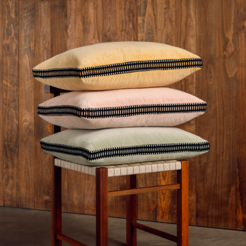 Drie Elitis Athena kussens in nieuwe kleuren liggen gestapeld op een houten stoel met geweven zitting voor een houten wand.