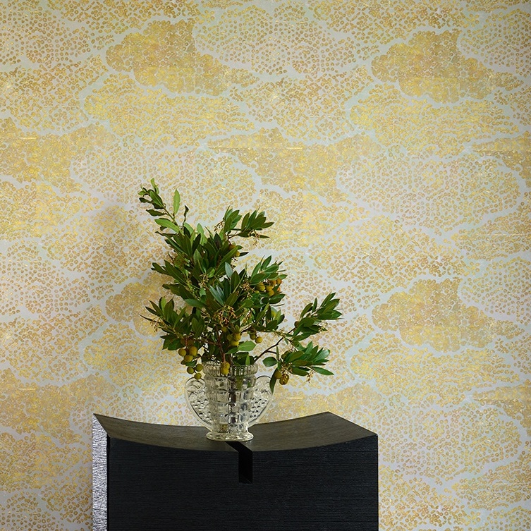Glazen vaas met groene takken op een zwart meubel, tegen een decoratieve gouden Elitis Bienfaisante Meditation-behangachtergrond.