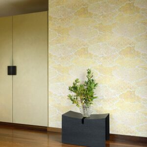 Moderne interieurhoek met Elitis Bienfaisante Meditation behang in zachte geel- en crèmetinten en een glazen vaas met groene takken op een zwart bijzettafeltje.