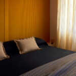 Slaapkamer met Elitis Colonne RM 1044 22 Rayures Jumelles behang achter een bed met donkere sprei en beige kussens.