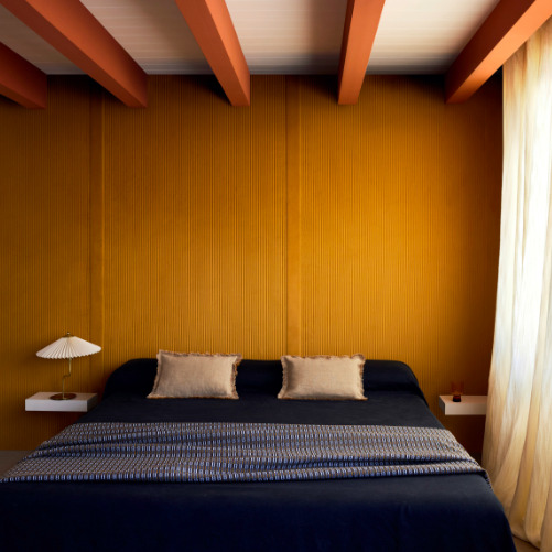 Slaapkamer met Elitis Colonne RM 1044 22 Rayures Jumelles behang in okergeel achter een donkerblauw bed met twee beige kussens.