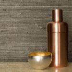 Glanzende metallic kom en koperen flesje staan voor Elitis Ecrin Faste behang met een luxe geweven textuur.