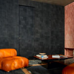 Moderne woonkamer met donker Elitis Empreintes RM 1040 80 behang uit de Marqueteries collectie, een leren oranje fauteuil en een lage houten salontafel.