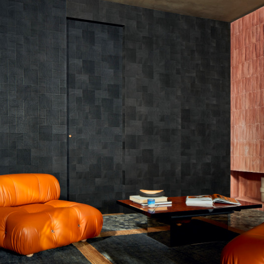 Moderne woonkamer met donker Elitis Empreintes RM 1040 80 behang uit de Marqueteries collectie, een leren oranje fauteuil en een lage houten salontafel.