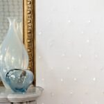 Glazen vazen op een marmeren tafel voor een gouden spiegel en een witte Elitis Ercin Naha-muur met subtiele structuur.