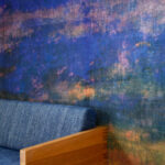 Detail van het Elitis Eternité VP 965 01 Atelier d'Artiste behang met een abstracte, kleurrijke textuur achter een houten bank met blauw gestoffeerde zitting.
