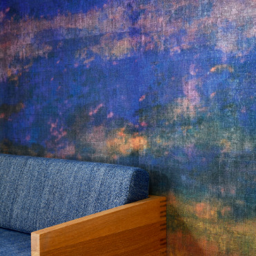 Detail van het Elitis Eternité VP 965 01 Atelier d'Artiste behang met een abstracte, kleurrijke textuur achter een houten bank met blauw gestoffeerde zitting.