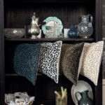 Vier Elitis Fauve-kussens met panterprint liggen op een donkere houten plank tussen decoratieve objecten en servies.