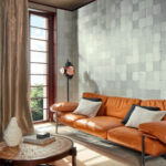 Moderne woonkamer met Elitis Feuilles d'or RM 1041 42 behang uit de Marqueteries collectie, een cognackleurige leren bank en grote raampartij.