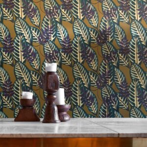 Behang van Elitis Flower Power Alocasia met groot tropisch bladmotief in groentinten en paarstinten op een okergele achtergrond, achter houten kandelaars op een marmeren tafel.