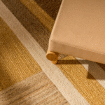 Detail van het Elitis Gustave TA 137 vloerkleed in okertinten met een beige meubelstuk op houten poot.