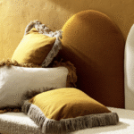 Stapel Elitis Honore kussens in okergeel en crème met franjerand op een zacht bekleed bankje tegen een okergele muur.